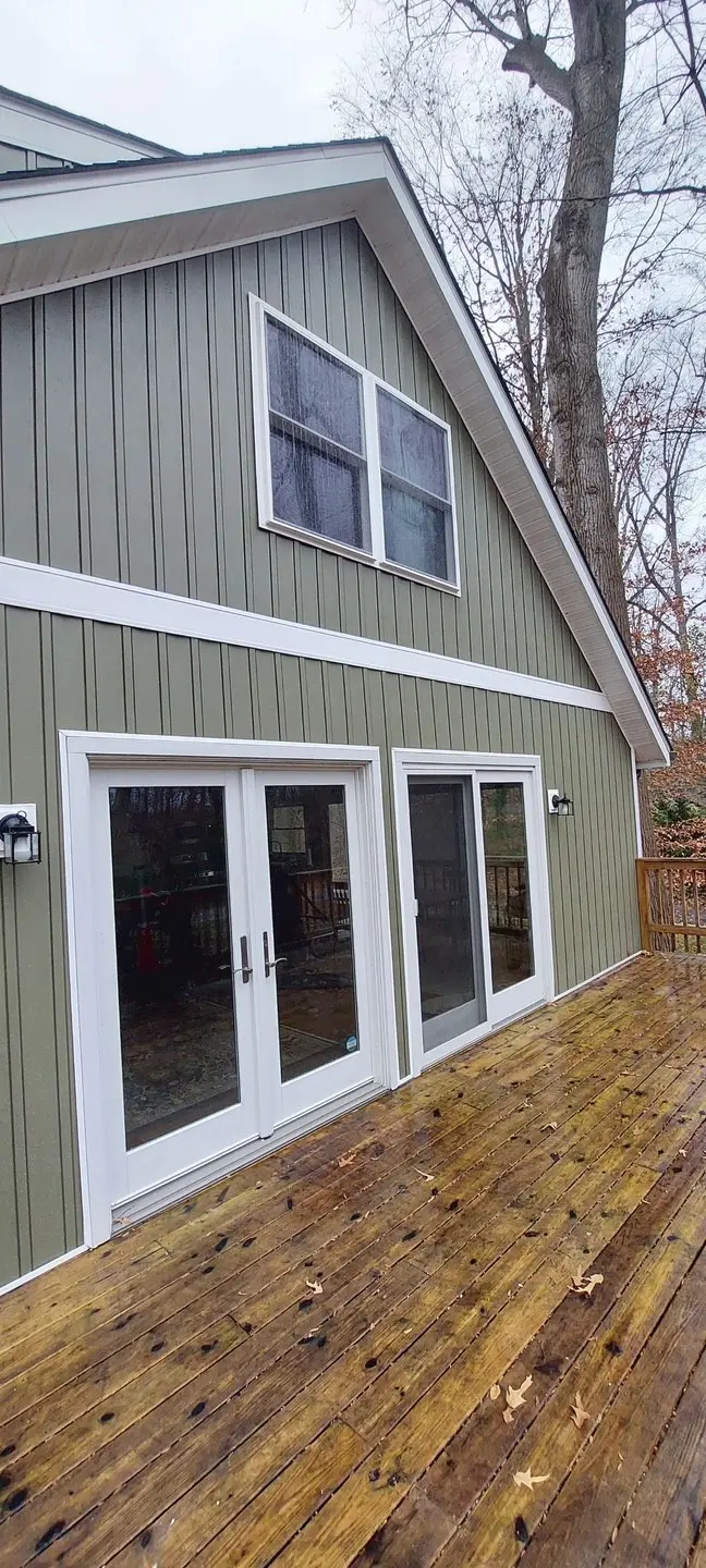 A renovated house exterior featuring sage green vertical board and batten siding, crisp white trim, and a set of white French doors opening onto a wet wooden deck.