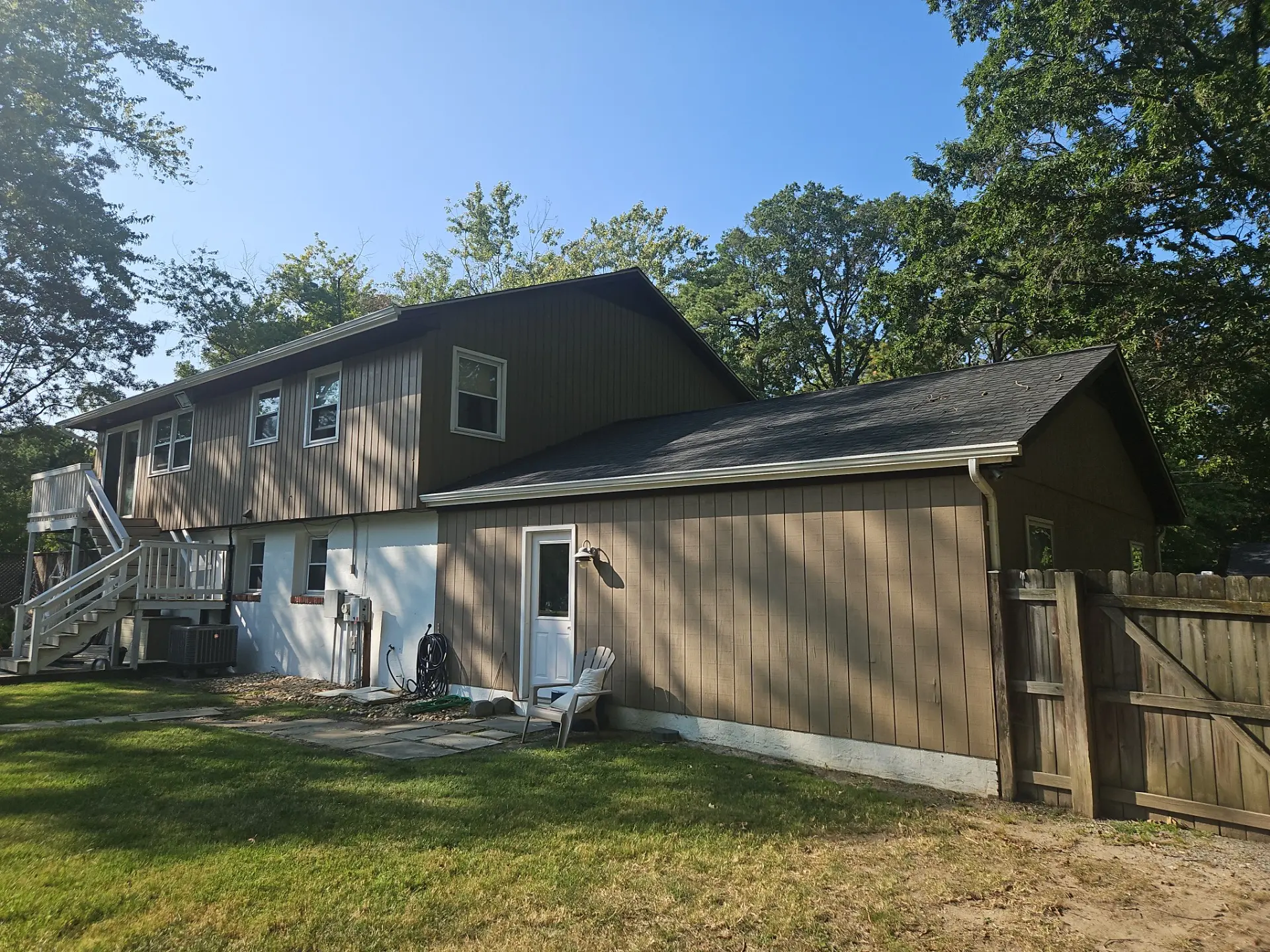 Two-story brown house with a deck and stairs, surrounded by greenery. Features include multiple windows and a fenced yard, ideal for outdoor living.