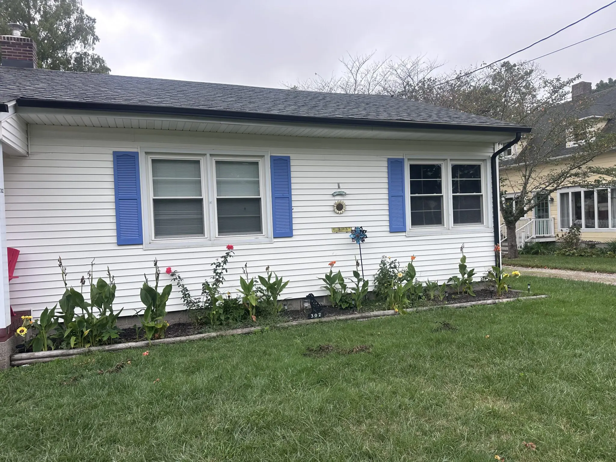 White house with blue shutters, featuring a flower garden in front. The scene captures a cozy residential setting with a cloudy sky.