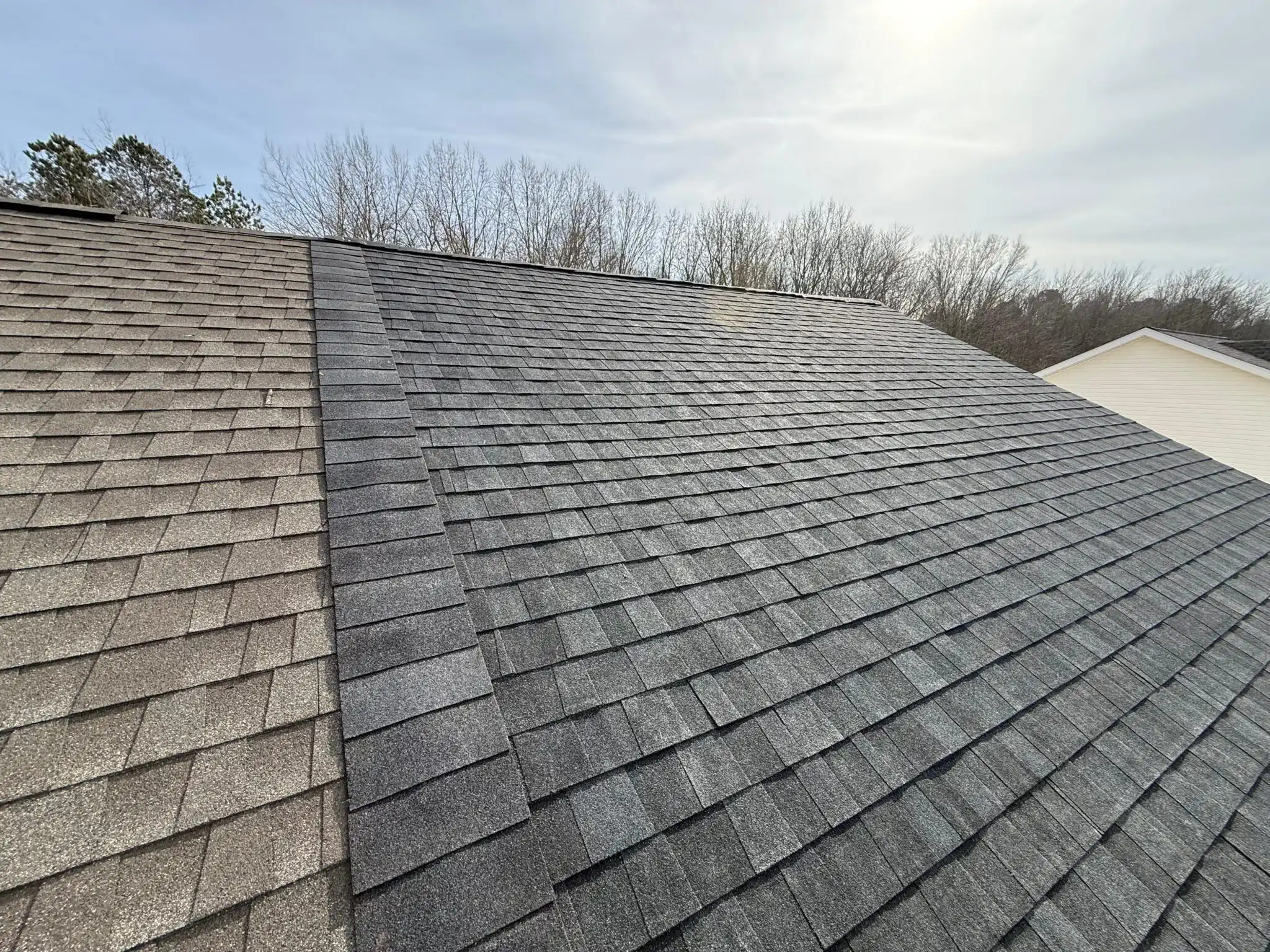 Lightly worn asphalt shingles on a sloped roof, showcasing a color transition from gray to dark gray, with sparse trees in the background.