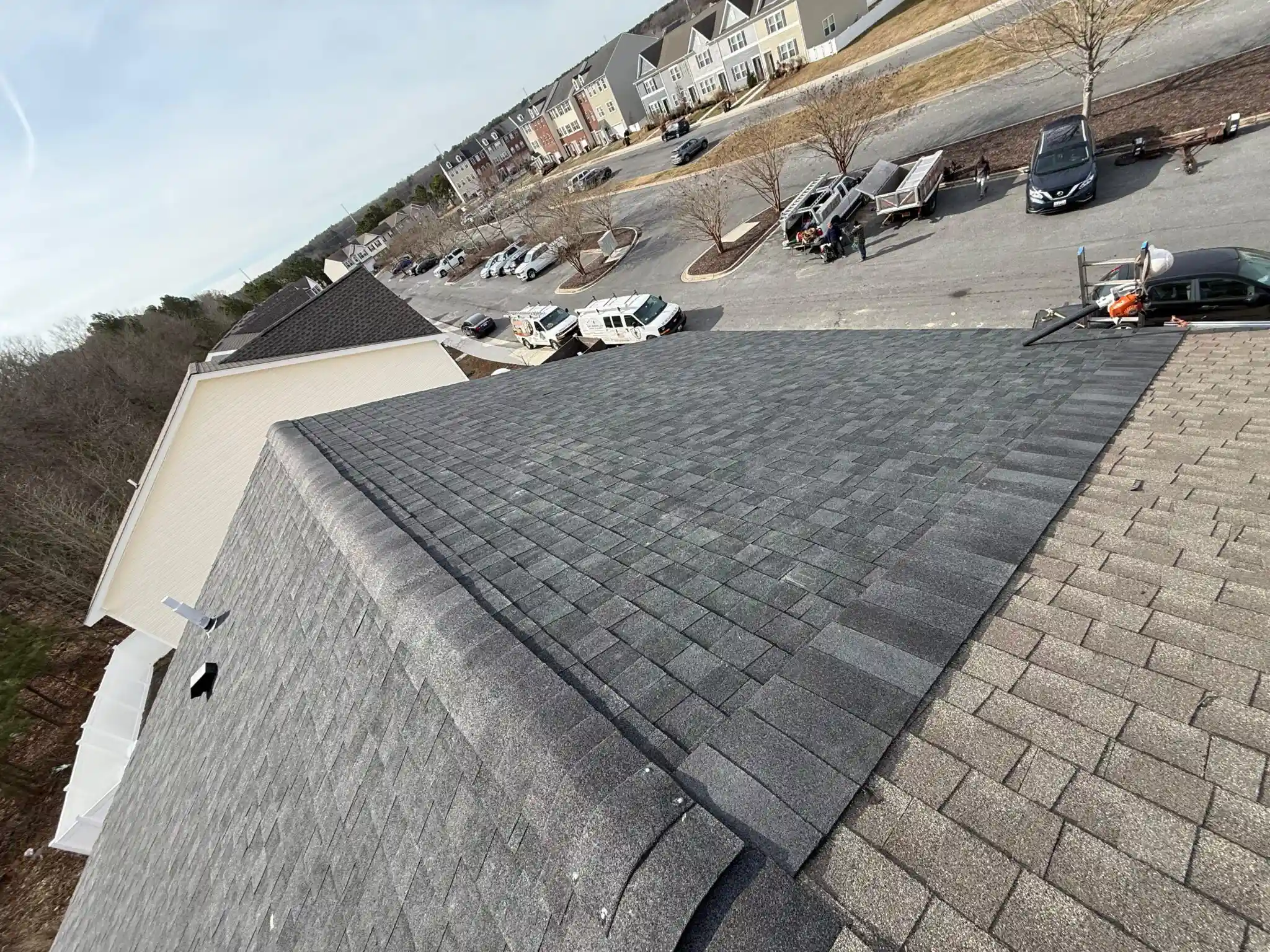 Sloped roof covered with dark gray shingles, showing adjacent rooftops and a residential area below. Construction vehicles are parked nearby.