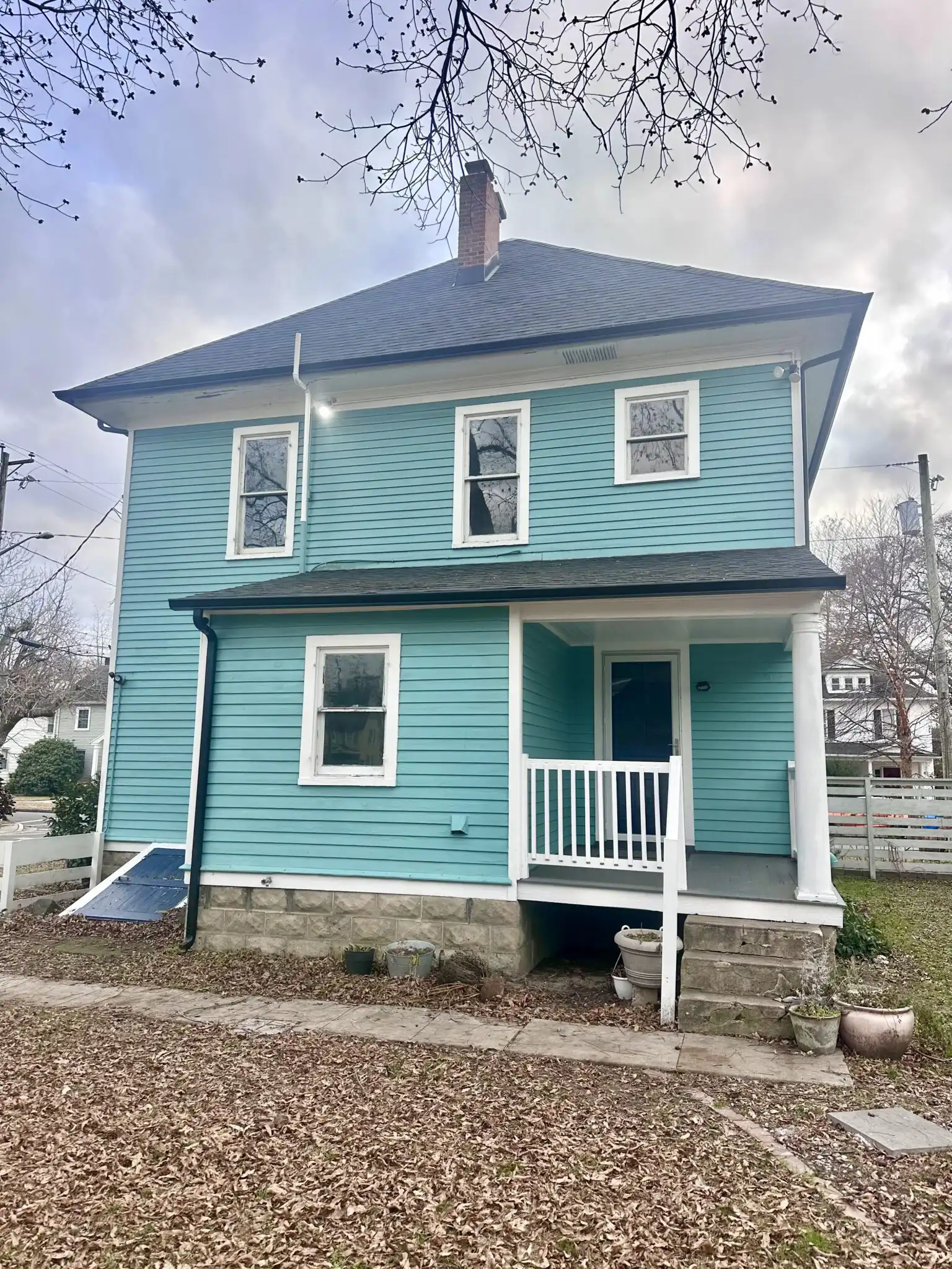 A two-story turquoise house with white trim and a front porch, surrounded by autumn leaves, under a cloudy sky. The image highlights residential architecture.