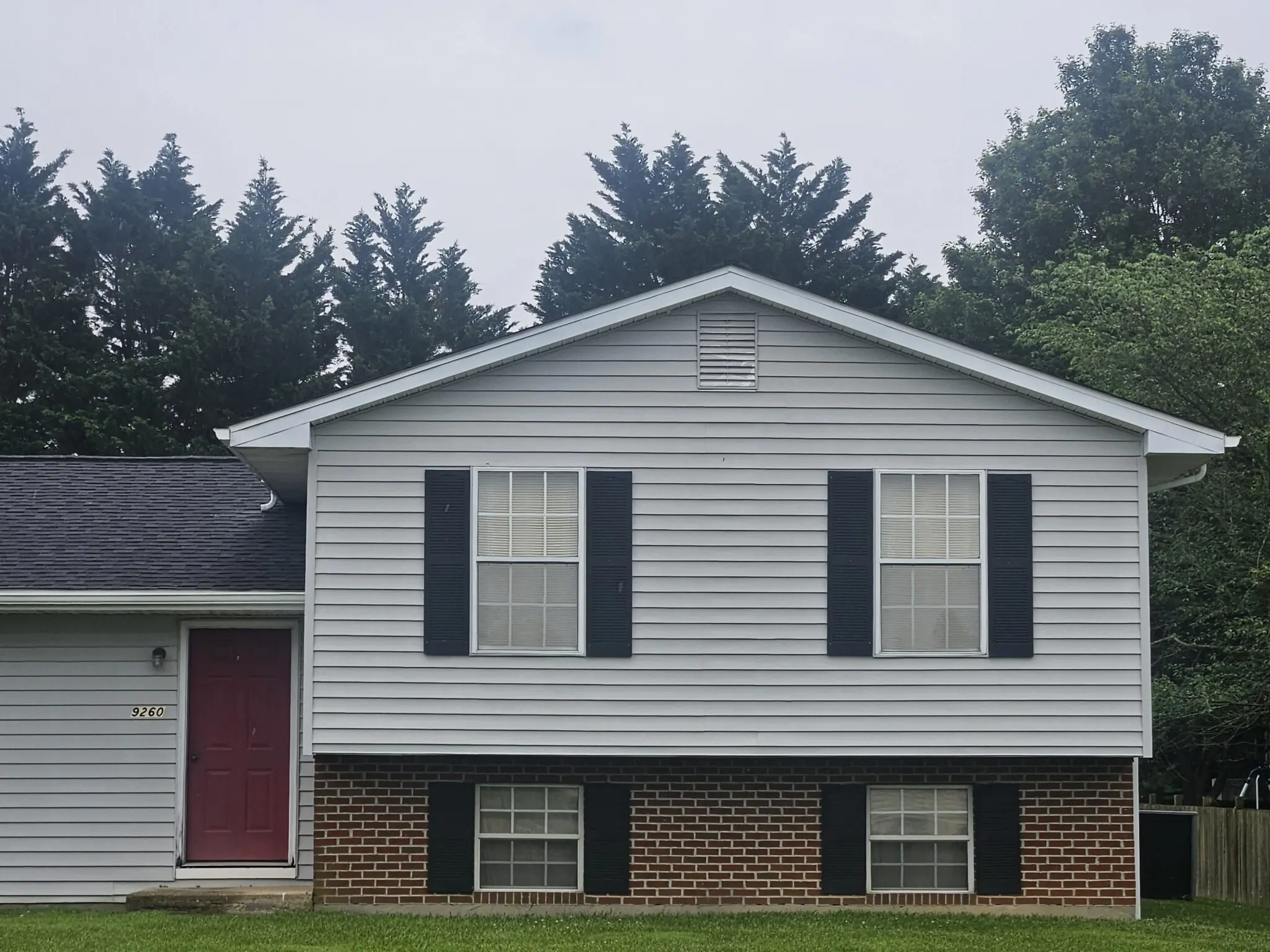 Gray house with a red front door, black shutters, and brick foundation, surrounded by tall trees. Captures residential architecture and exterior design.