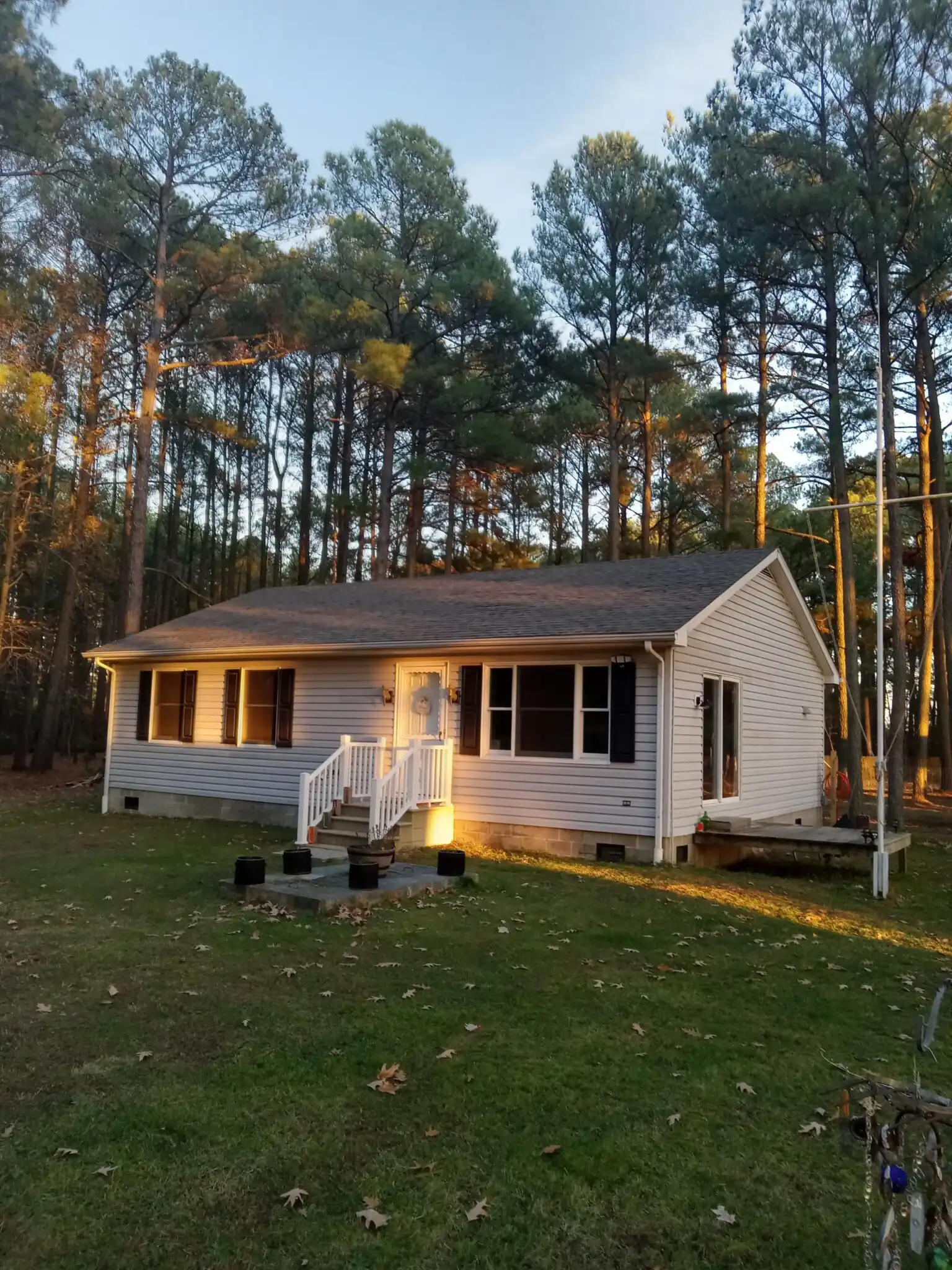 Single-story house surrounded by tall trees, featuring a front porch with a white railing and landscaped yard. Evening light highlights its exterior.