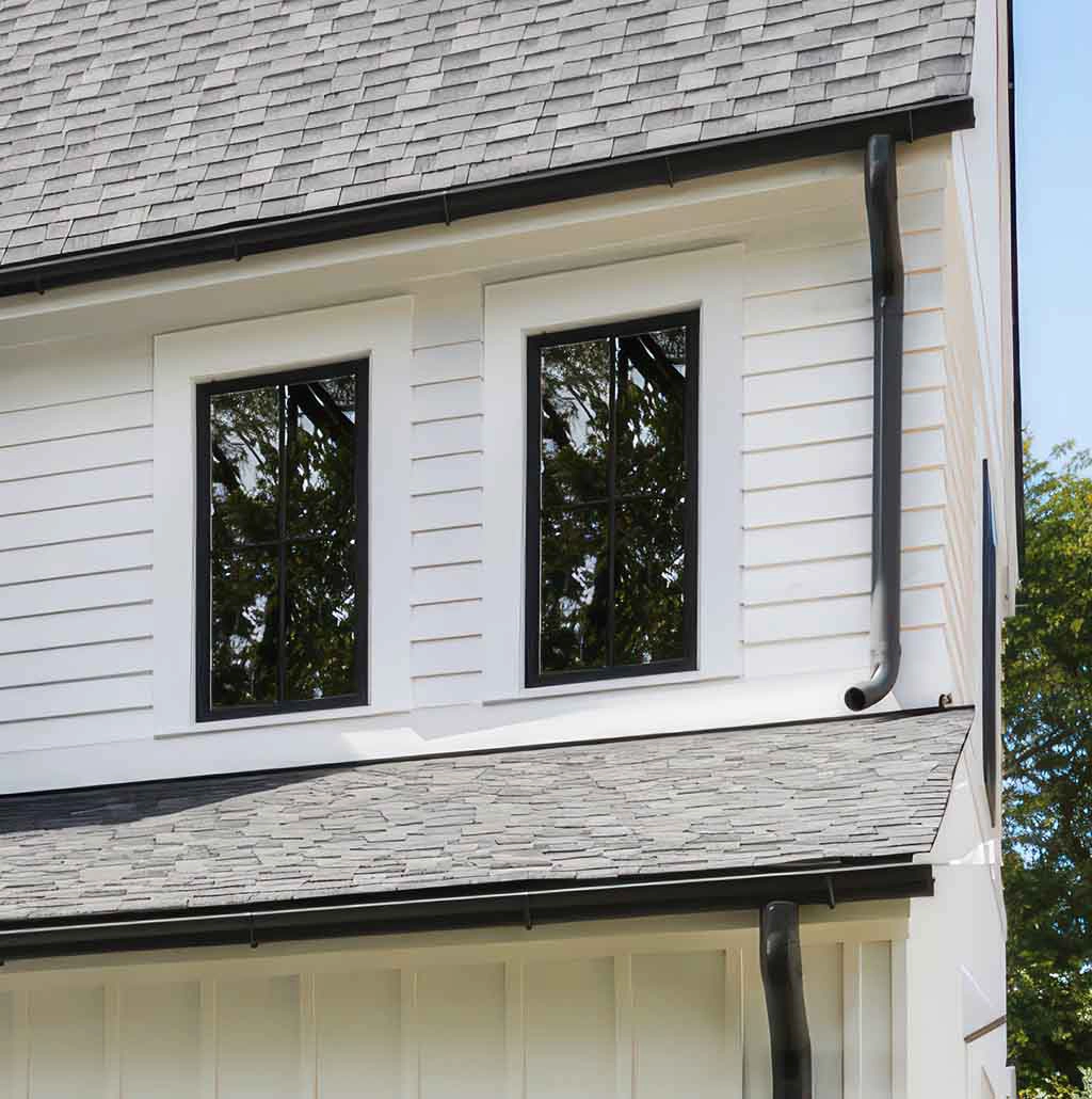 Two black-framed windows on a contemporary house, reflecting surrounding trees. The clean white siding and gray shingle roof emphasize modern architecture.