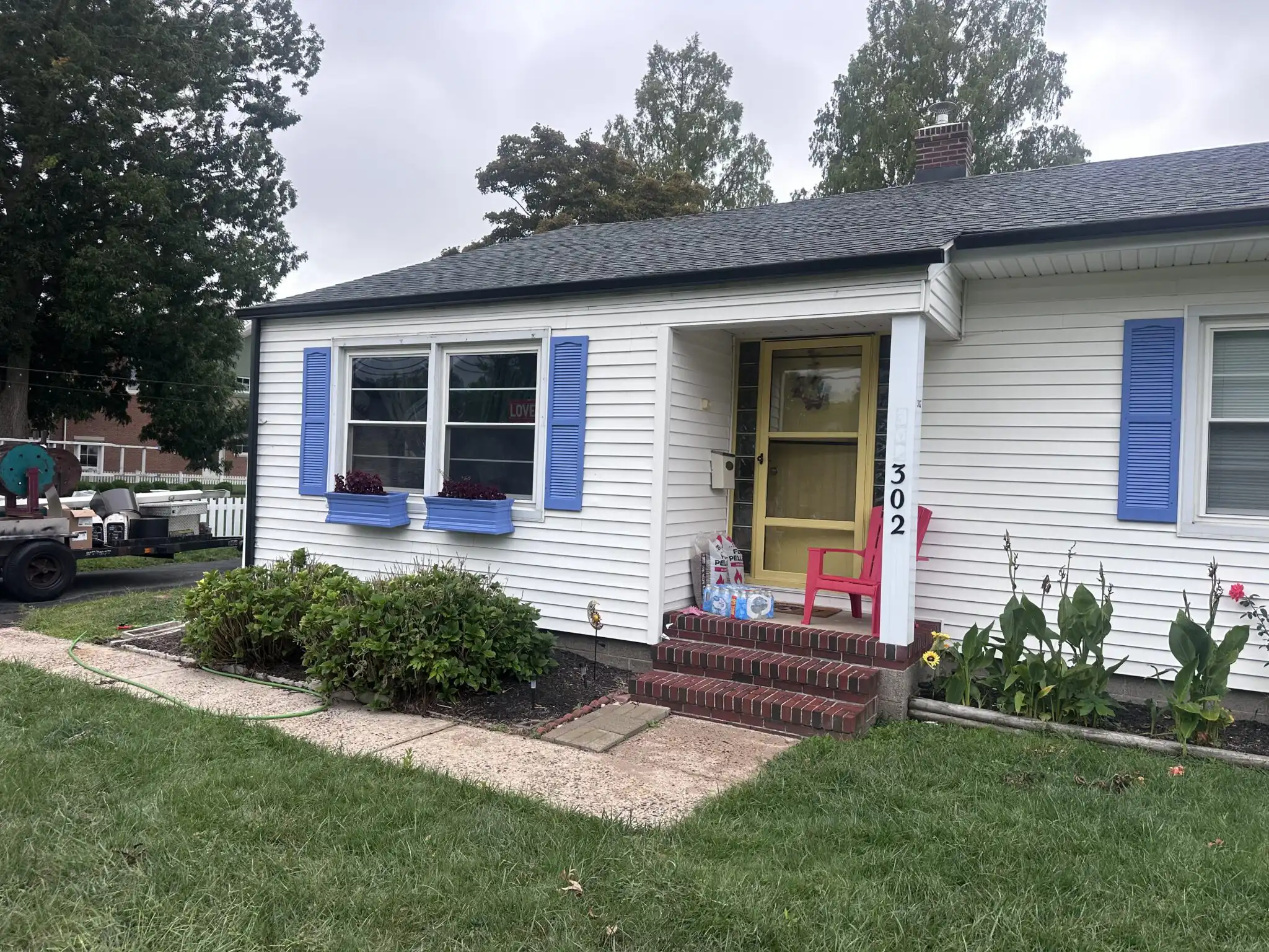 Charming white house with blue shutters and a yellow front door. Features a brick porch, flower boxes, and a pink chair on the steps.